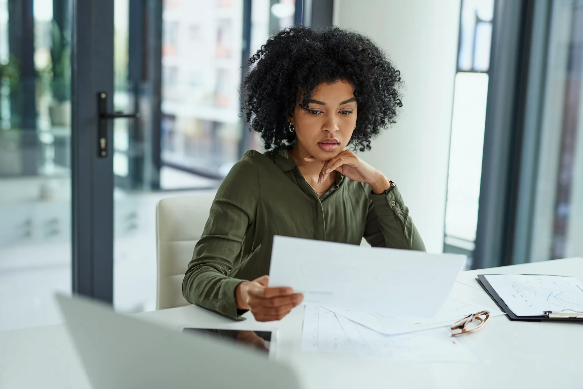 African-American woman reviews paperwork from her insurance settlement at a desk in front of glass doors.