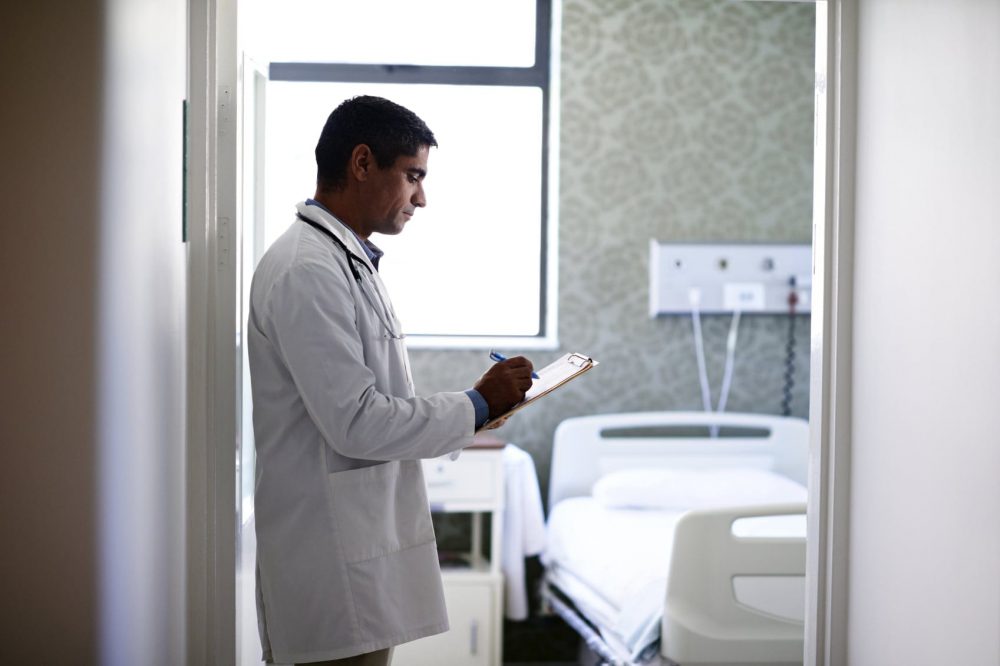Male Doctor Completing Medical Forms In Hospital Room Stock Photo