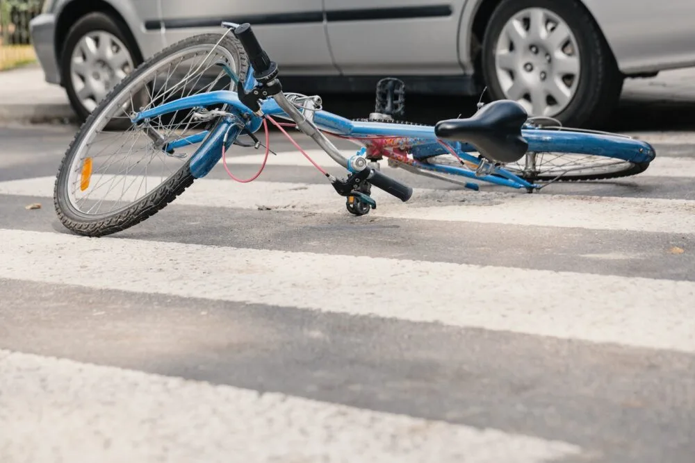 A blue bicycle lies in the middle of the street beside a gray sedan.