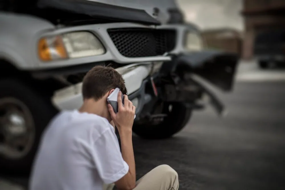 A man is sitting next to a damaged vehicle while talking on the phone.