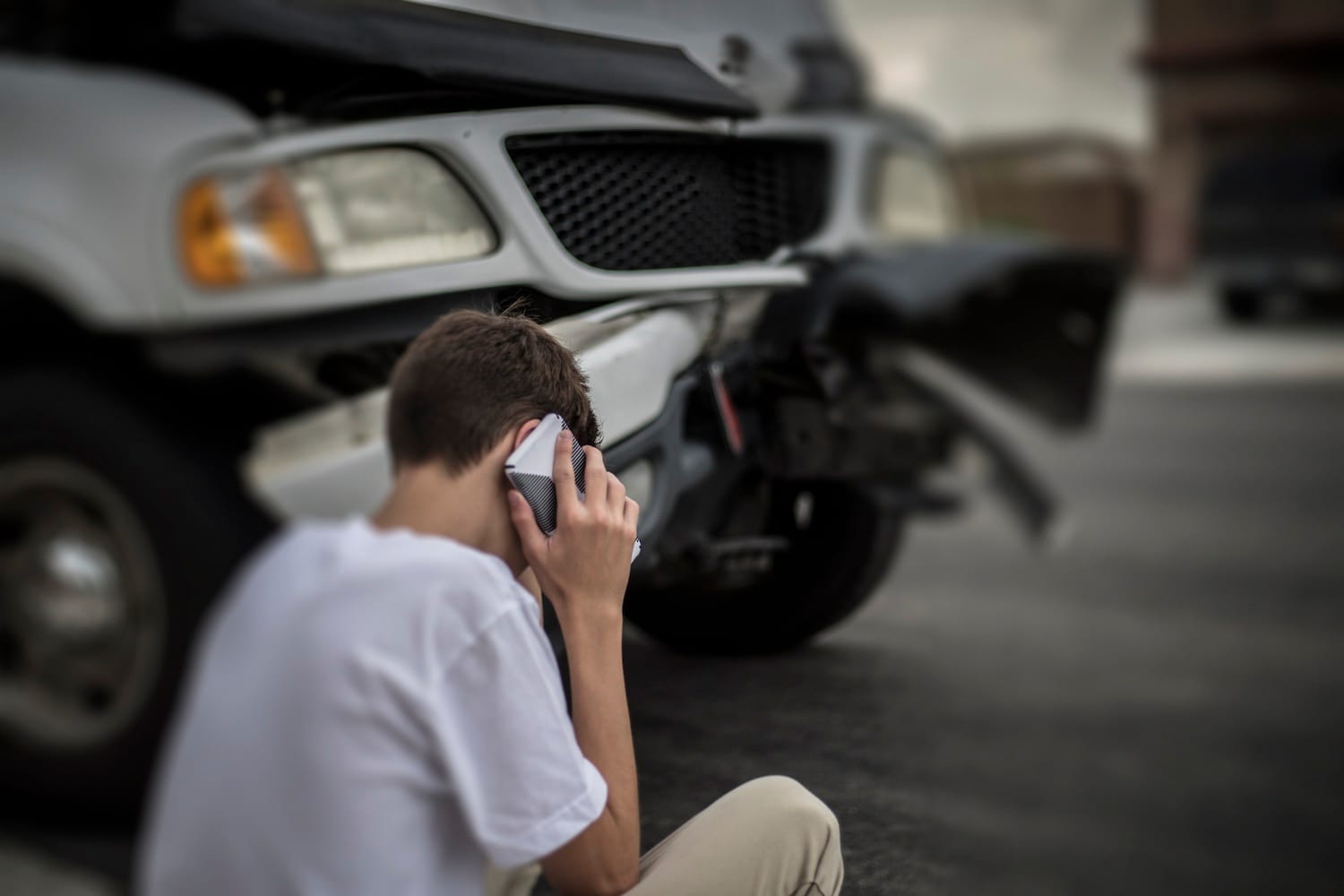 A man is sitting next to a damaged vehicle while talking on the phone.