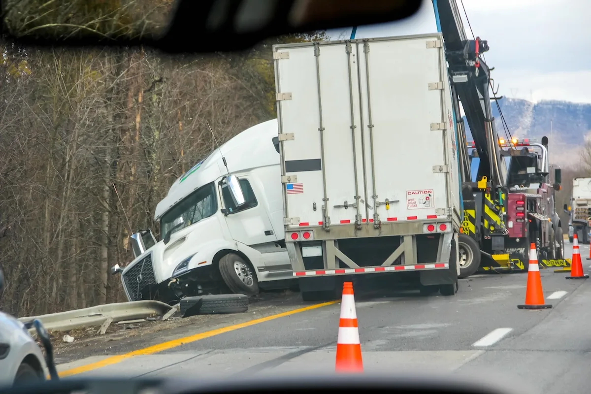 A wrecked semi-truck leaning off the side of the road. The trailer is positioned normally, but the cab is turned around to the left of the trailer at an angle. Cones are set up on the road while traffic passes by.