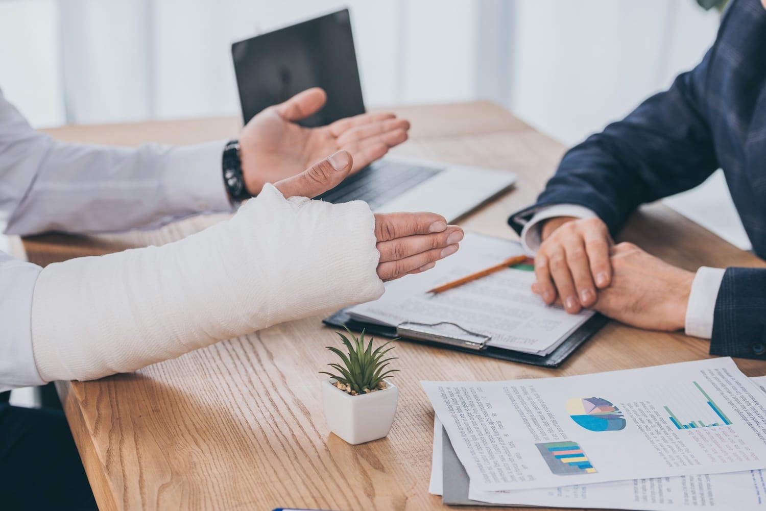 A person with their arm in a cast is speaking to a lawyer at a desk with papers, a small plant, and a computer also on the desk.