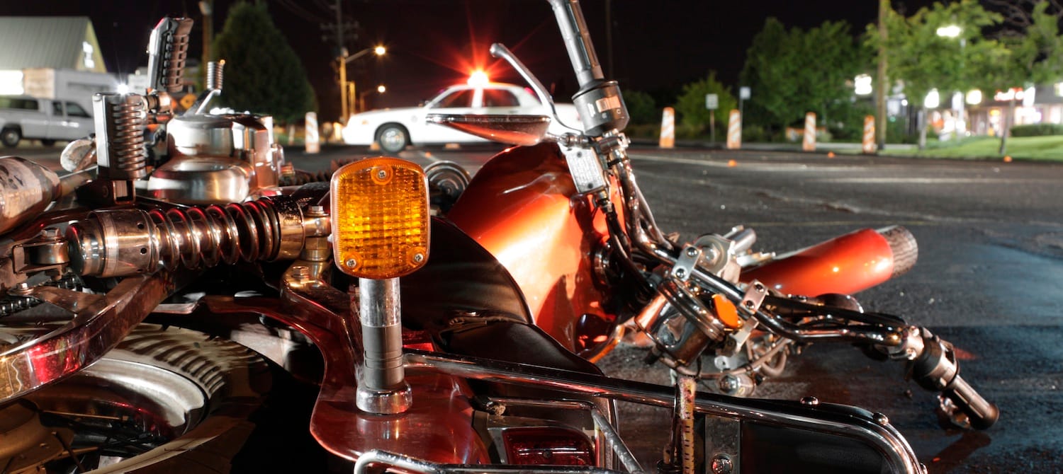 Close-up of a damaged motorcycle lying on the road at night after an accident, with a police car in the background flashing its lights.