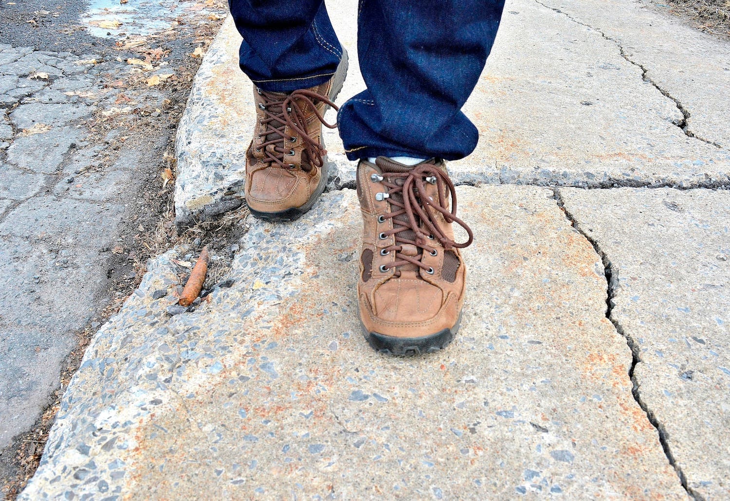 Someone walking on a broken sidewalk in brown shoes and blue jeans.