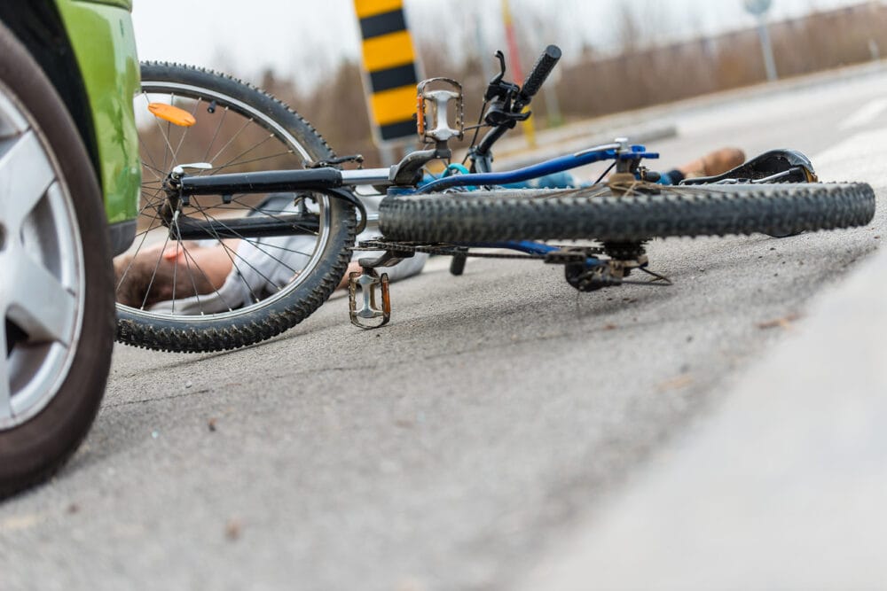 A crashed bicycle rider on the road in front of a green car.