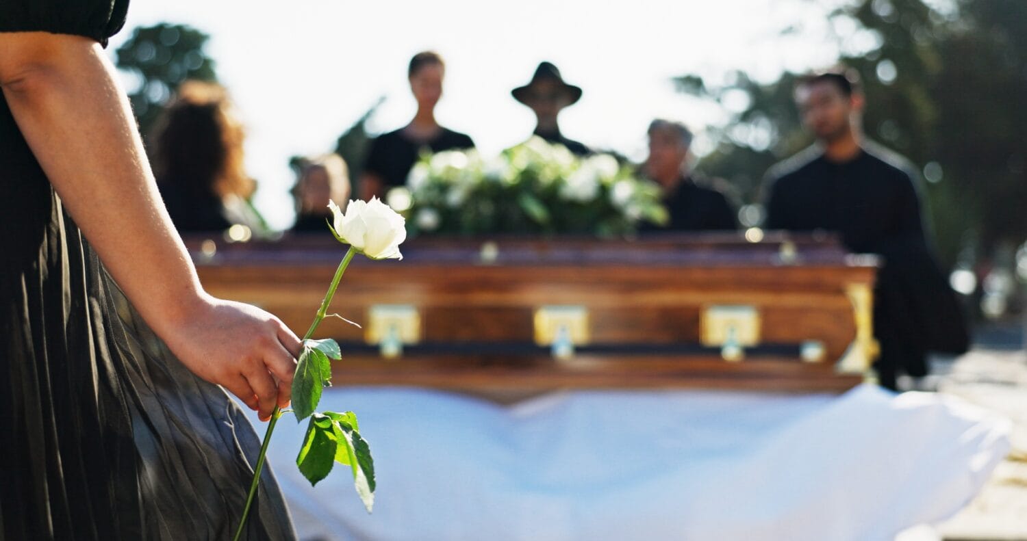 A close-up of a woman holding a white rose at a funeral, standing in front of a coffin. Other family members stand behind the coffin.