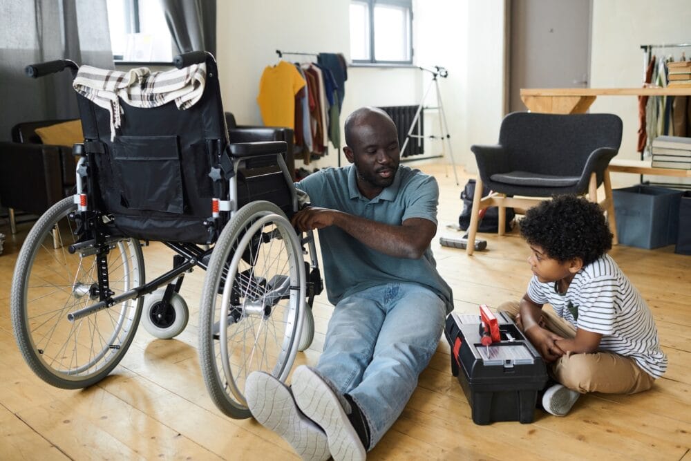 Disabled man sits on the floor next to his son while leaning against a wheelchair.
