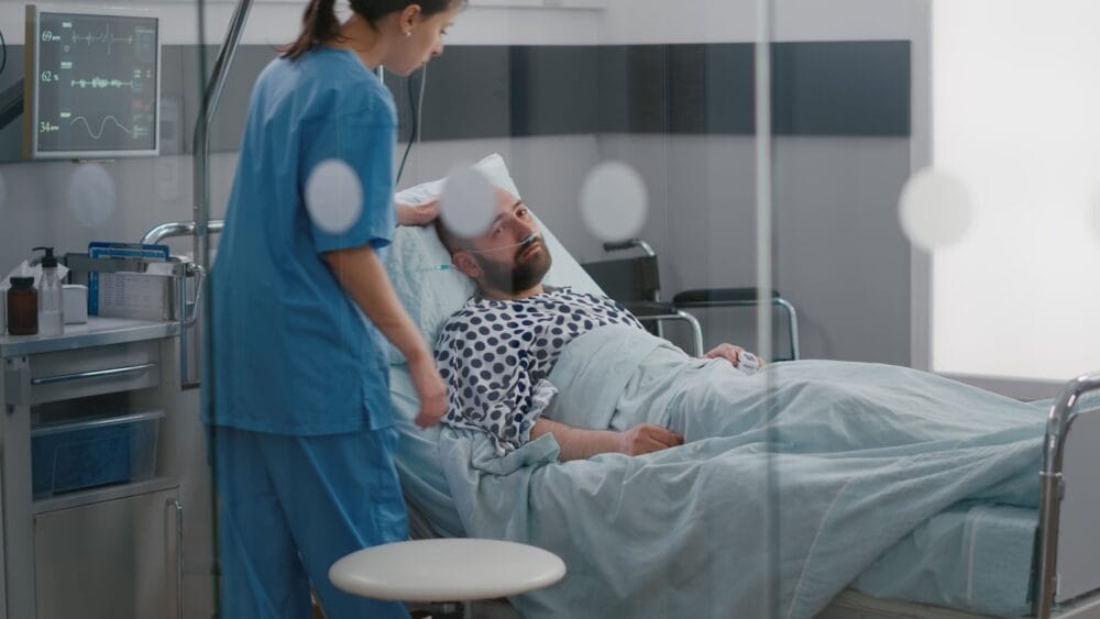 Sick man in a hospital bed wearing a nasal respirator as a nurse stands by his bed.