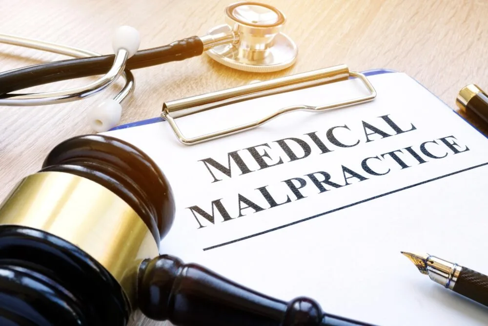A gavel and stethoscope lie on a table next to a pen and clipboard. The paper on the clipboard reads “MEDICAL MALPRACTICE”.