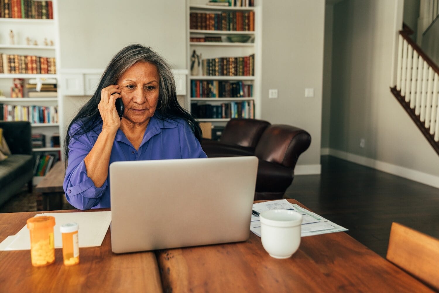 Woman talks on phone while viewing laptop with prescription bottles next to her.