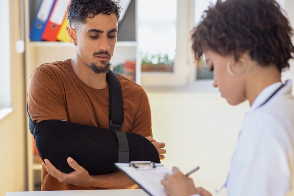 A man with his injured arm in a sling speaks with a medical provider who is taking notes on a clipboard.