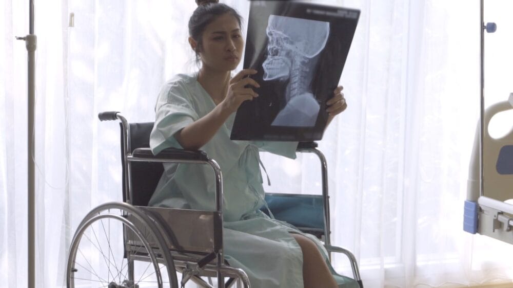 Woman in wheelchair examines head X-ray while sitting next to hospital bed.