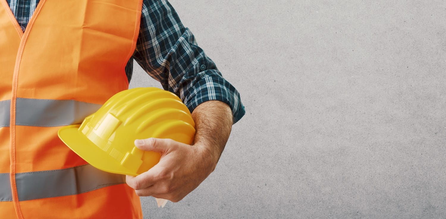 A man slightly out of frame is holding a protective helmet and wearing an orange safety vest.