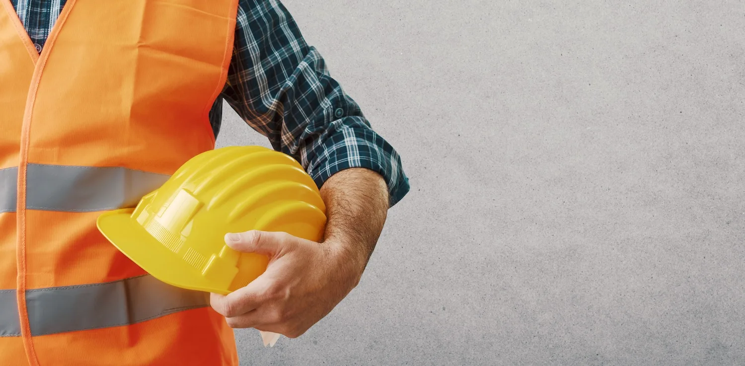 A man slightly out of frame is holding a protective helmet and wearing an orange safety vest.