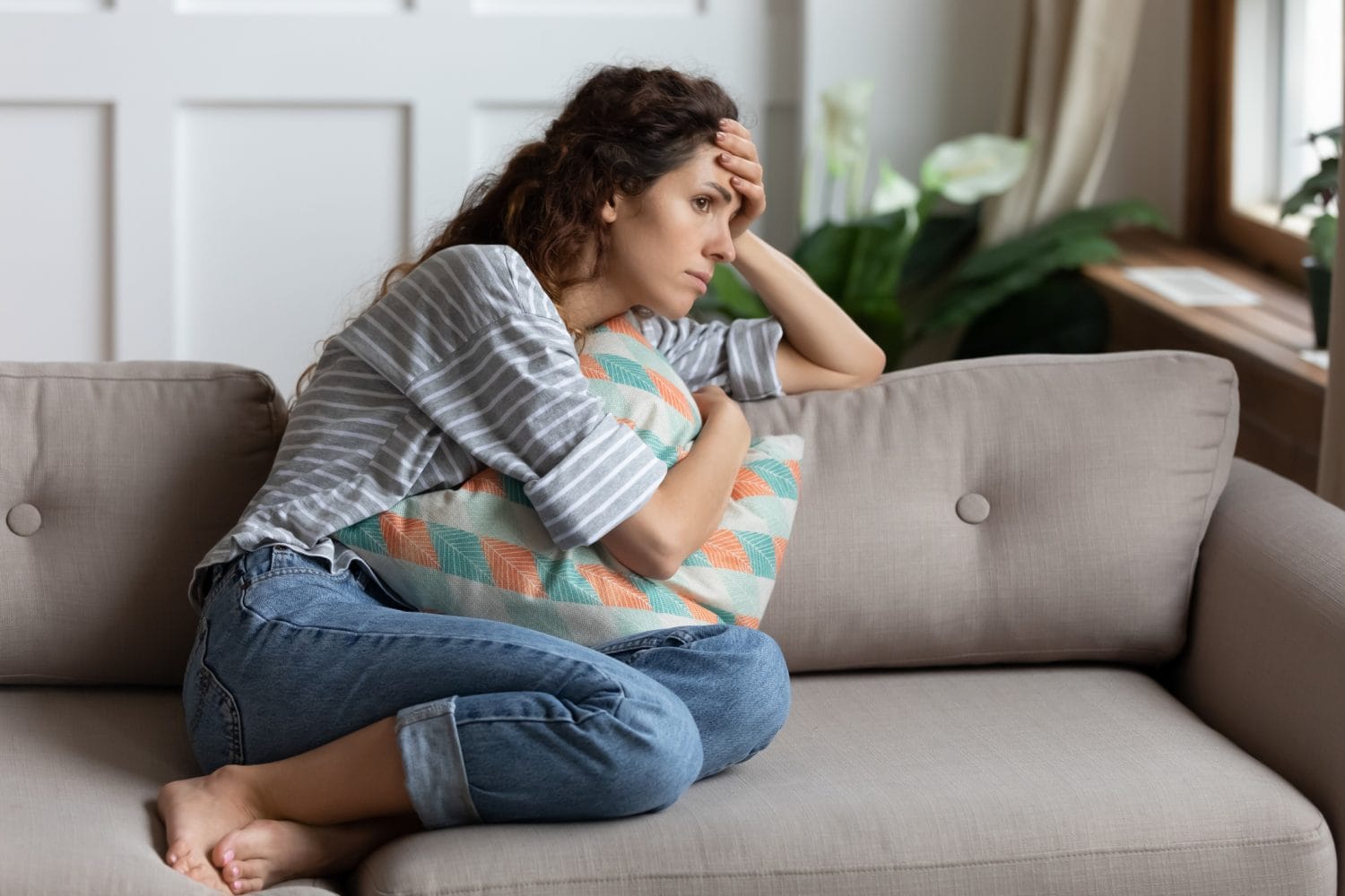 An unhappy woman wearing jeans sits on a couch with her legs folded while hugging a pillow.