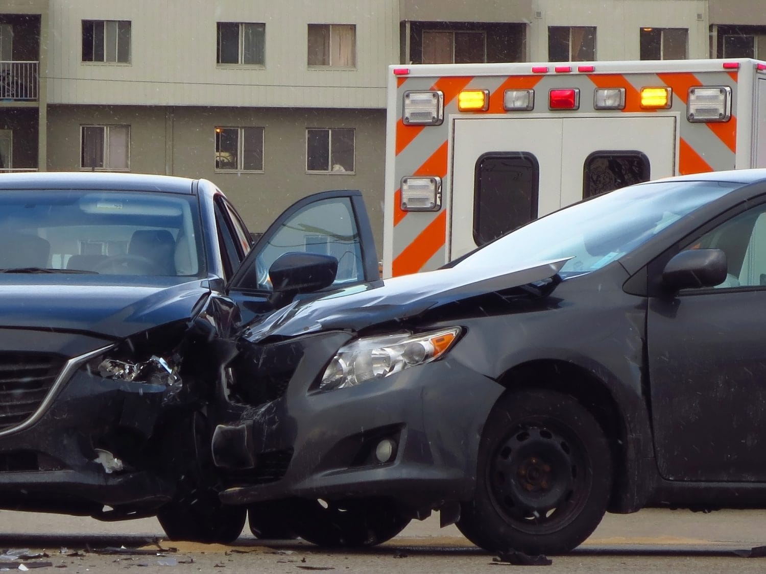 Two dark sedans involved in a front-end collision sit in front of an ambulance parked near an apartment complex.