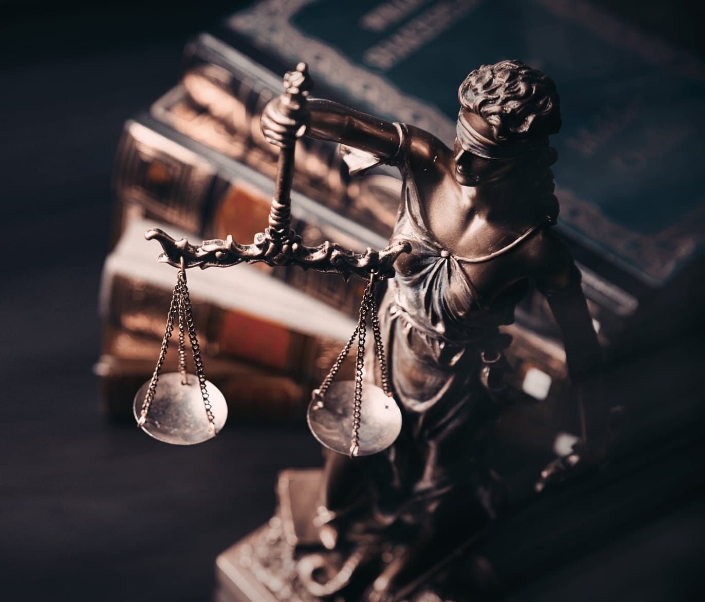 A bronze statue of Lady Justice holding scales stands in front of a stack of historic legal books.