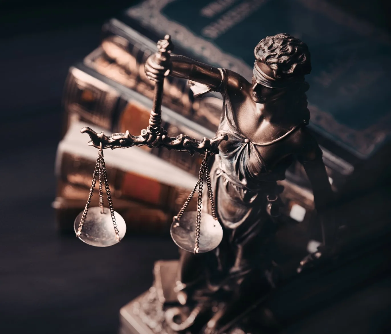 A bronze statue of Lady Justice holding scales stands in front of a stack of historic legal books.