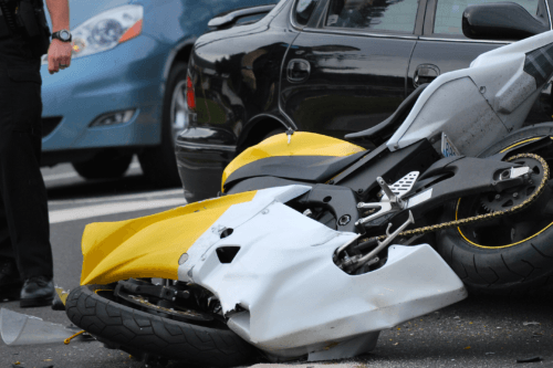 A yellow and white motorcycle crashed on the ground next to a black car. A man stands to the side, and another car can be seen behind him.